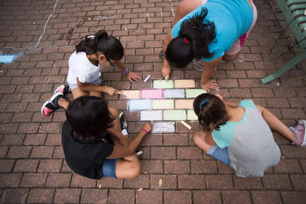 Four girls use chalk to turn make a rainbow brick road.