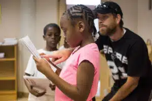 Trinity Gilchrist, 8, center, reads a story she wrote while her teacher, Jason Harrison, right, helps her during The Serna Summer Enrichment Program. WENDI POOLE / SPECIAL TO FOLO MEDIA